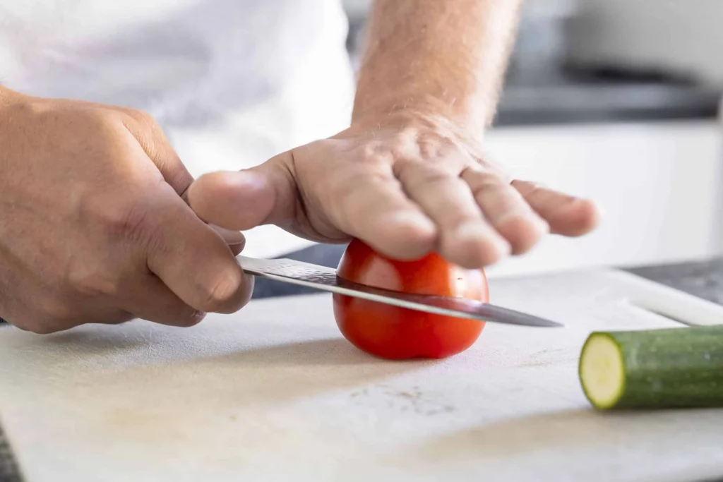 A man using a knife to slice a tomato on a cutting board.
