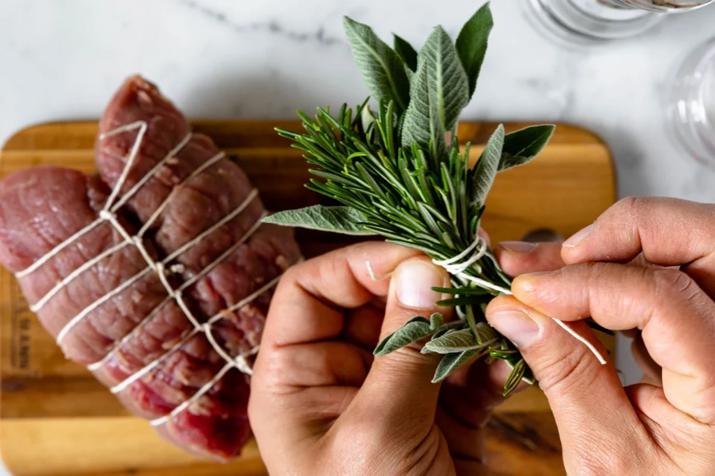 A person wrapping a piece of meat with sprigs of rosemary.