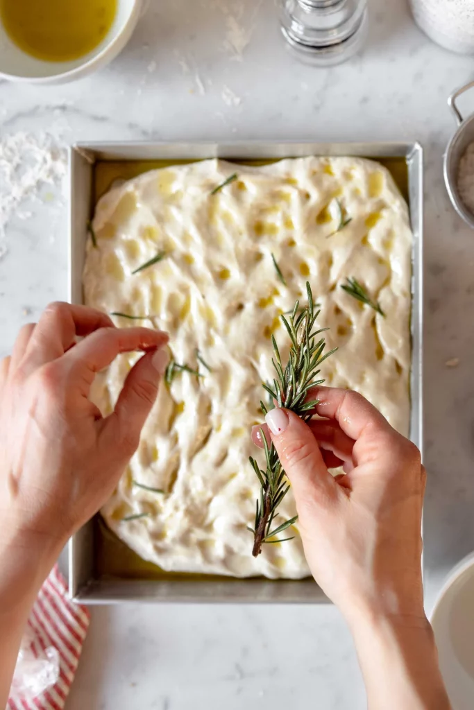 A person sprinkling rosemary on a pizza dough.