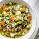 A bowl of soup with carrots, peas and parsley.