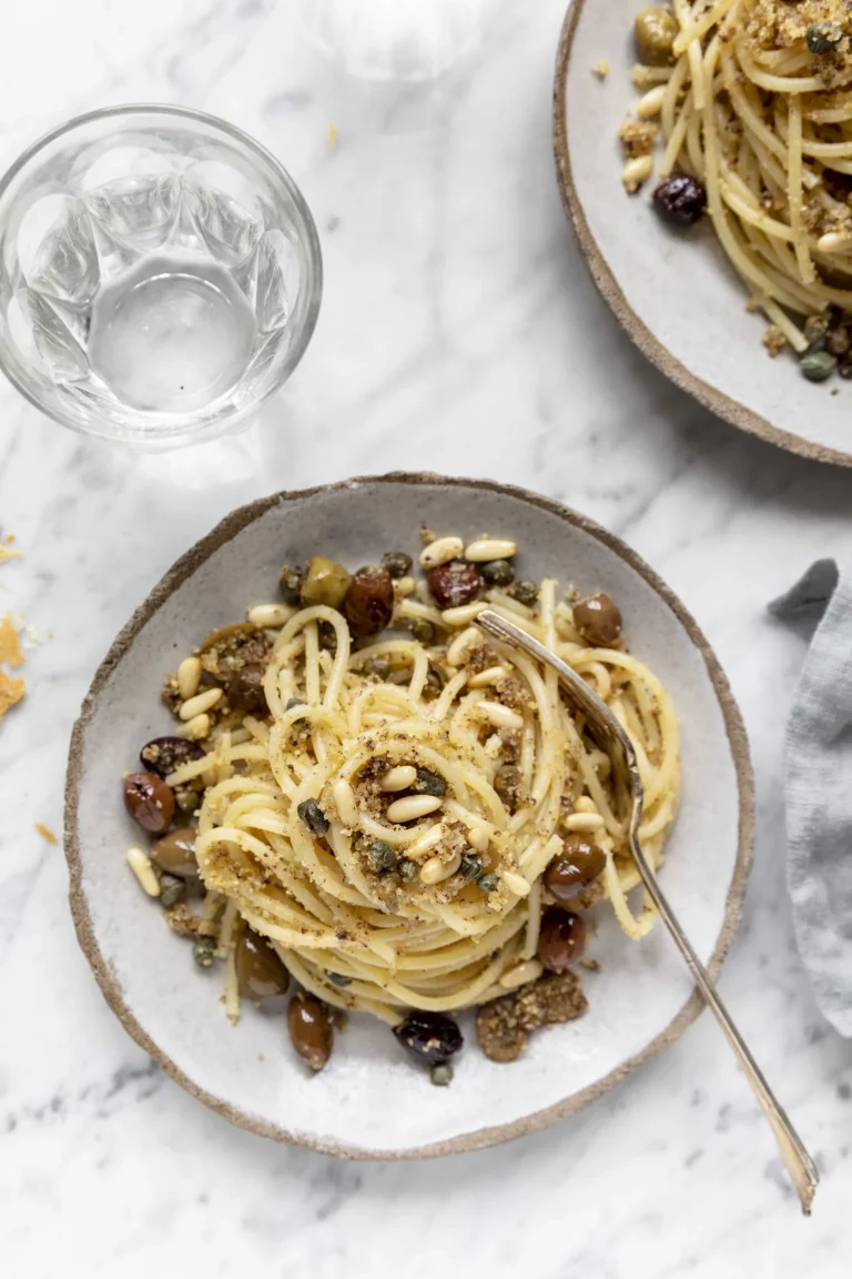 Two plates of spaghetti with nuts and olives on a marble table.
