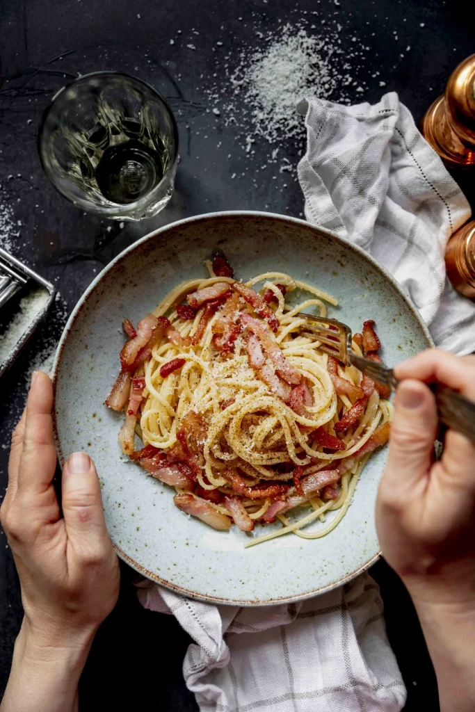 Hands holding a plate of spaghetti with bacon and parmesan cheese.