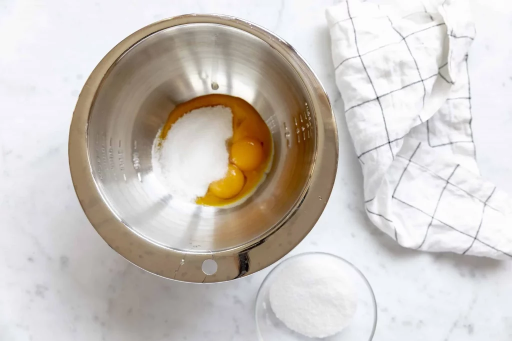 A mixing bowl with eggs and sugar next to it, perfect for making a delicious tenerina cake.