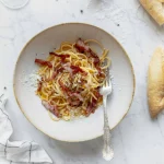 A bowl of pasta with bacon and bread on a marble table.