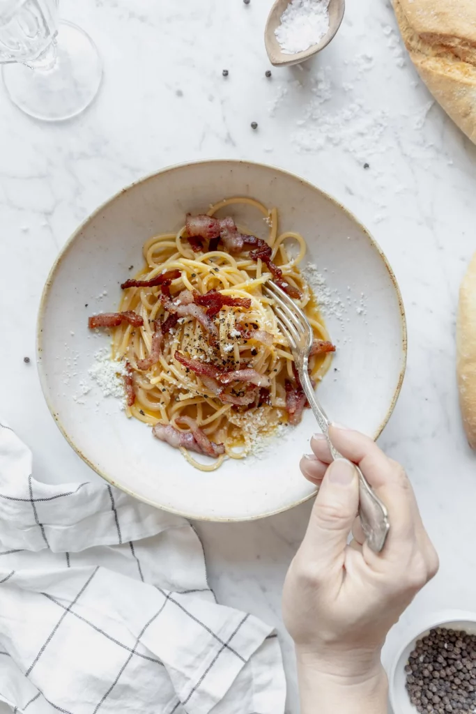 A plate of spaghetti with bacon and bread on a marble table.