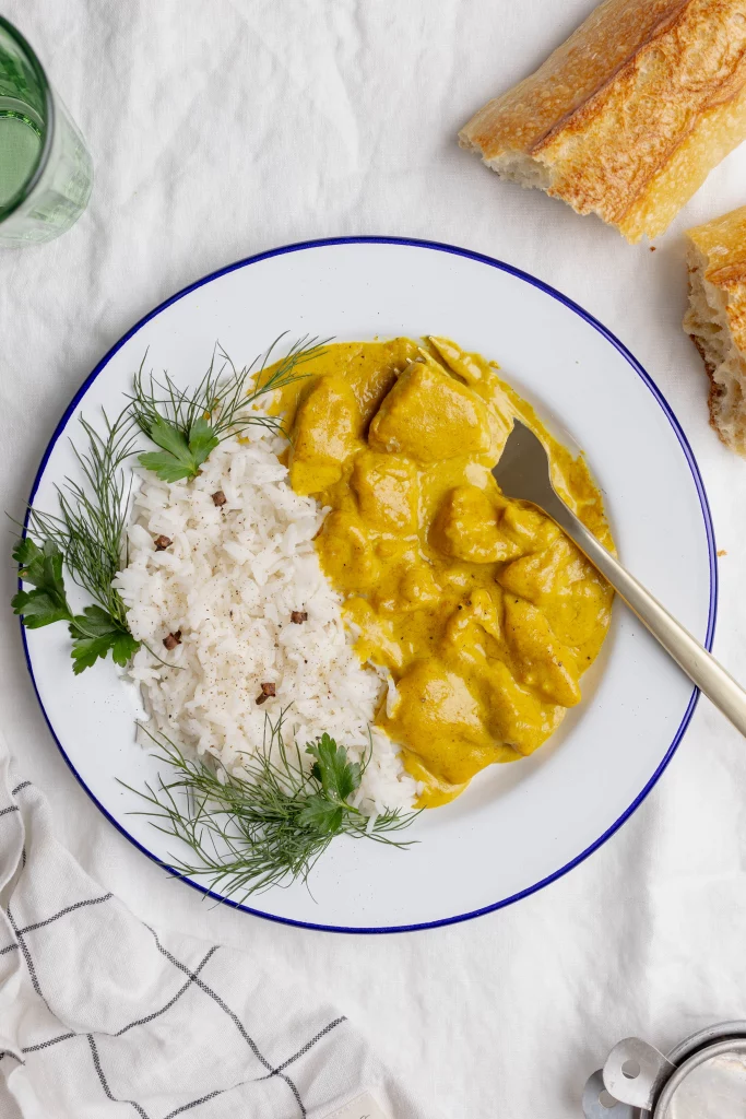 A plate of chicken curry with white rice garnished with fresh herbs, accompanied by a baguette on a white background.