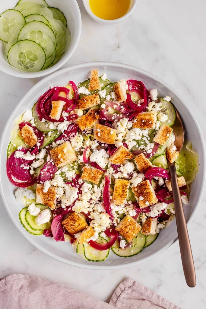 Greek salad with cucumbers, red onions, croutons, and feta cheese in a white bowl, with a fork and a side bowl of dressing.