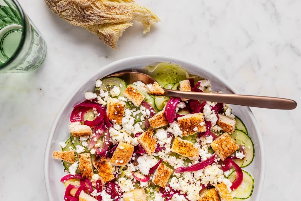 A bowl of salad with cucumbers, pickled onions, feta cheese, and croutons, accompanied by a glass of water and a piece of bread.