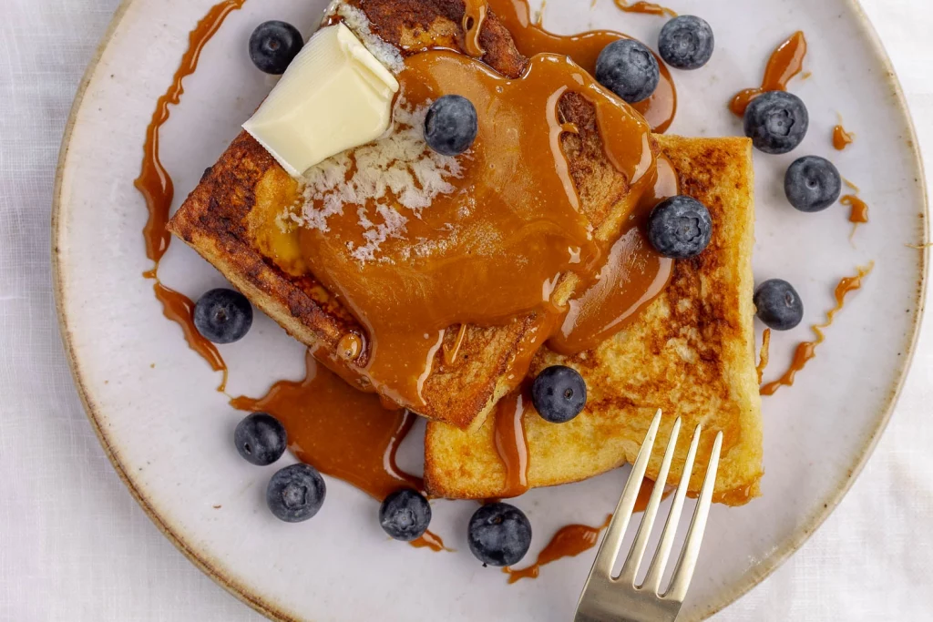 Plate of french toast topped with caramel sauce, a slice of butter, and fresh blueberries, with a fork on the side.