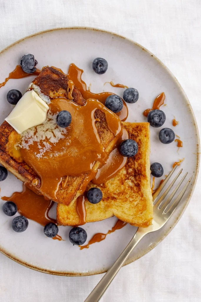French toast with caramel sauce, fresh blueberries, and a slice of butter, served on a white plate with a fork.