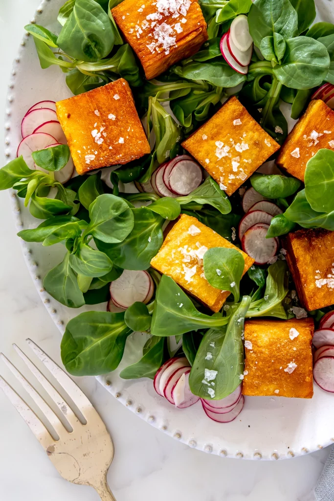 A plate of fresh salad topped with golden-brown Thousand Layers Potatoes cubes, thinly sliced radishes, and leafy greens, garnished with coarse salt, accompanied by a fork.