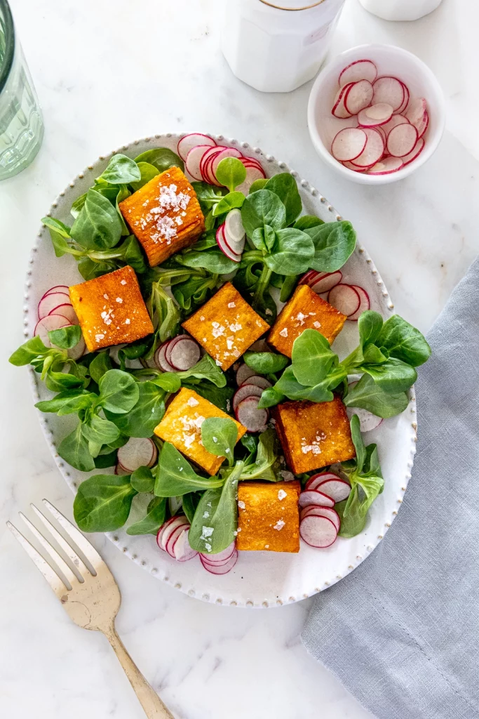 A fresh salad with spinach, sliced radishes, and golden brown Thousand Layers Potatoes, sprinkled with salt, served on a white plate with a fork beside it.