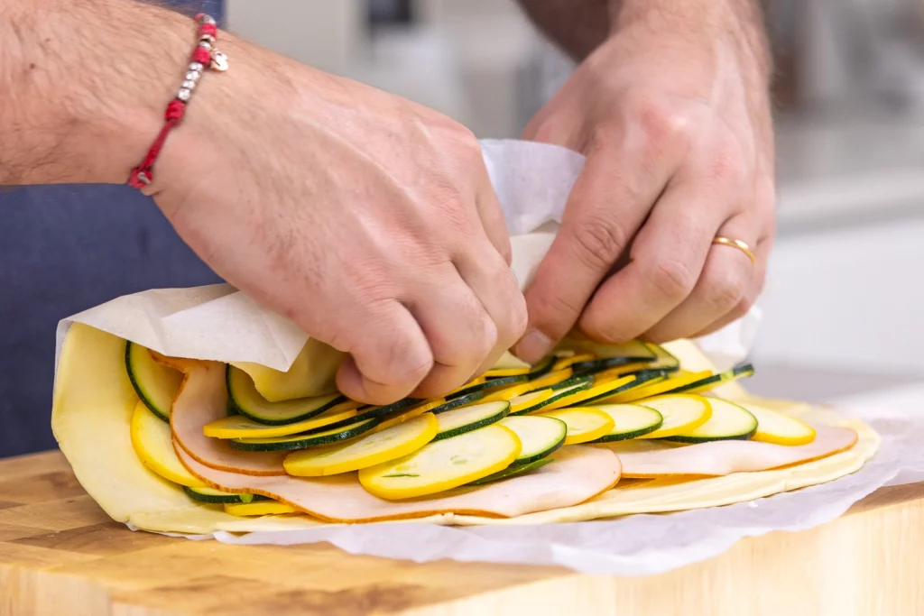 Hands assembling a roll with layers of turkey, zucchini, and yellow zucchini on parchment paper over a wooden cutting board.